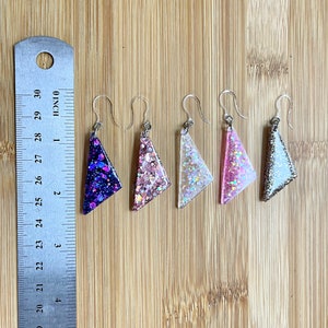 May include: Five pairs of triangle-shaped earrings with glitter embedded in the resin. The earrings are in shades of purple, pink, white, and silver.