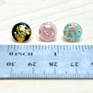 Puede incluir: Tres cabujones redondos de vidrio con detalles de pan de oro. Los cabujones son negros, rosas y azul claro. Los cabujones tienen un diámetro de aproximadamente 1,25 cm.