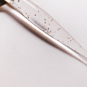 May include: A clear glass dropper with gold flecks suspended within the liquid. The dropper is angled diagonally across a white background, creating a minimalist aesthetic. The gold flecks add a touch of luxury to the design.