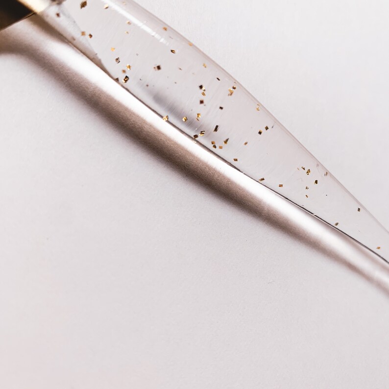 May include: A clear glass dropper with gold flecks suspended within the liquid. The dropper is angled diagonally across a white background, creating a minimalist aesthetic. The gold flecks add a touch of luxury to the design.