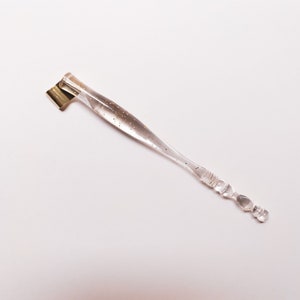 May include: A clear calligraphy pen with gold flecks and a gold nib. The pen has a clear, textured handle and is designed for detailed writing and drawing. The pen is angled for comfortable use.