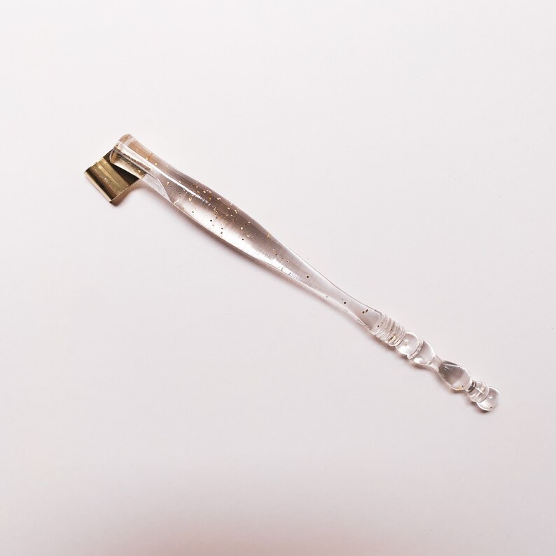 May include: A clear calligraphy pen with gold flecks and a gold nib. The pen has a clear, textured handle and is designed for detailed writing and drawing. The pen is angled for comfortable use.