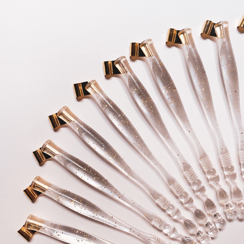 May include: A collection of clear calligraphy pen holders with gold-colored nibs and glitter accents. The pens are arranged in a fan shape against a white background, showcasing their elegant design. Ideal for calligraphy enthusiasts.