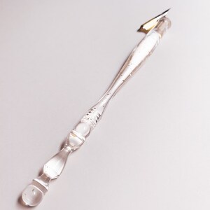 May include: A clear glass calligraphy pen with a gold nib. The pen's handle is transparent, with gold flecks and decorative bulbous sections. The pen is designed for writing and drawing.