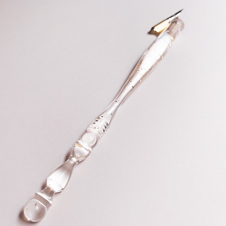 May include: A clear glass calligraphy pen with a gold nib. The pen's handle is transparent, with gold flecks and decorative bulbous sections. The pen is designed for writing and drawing.