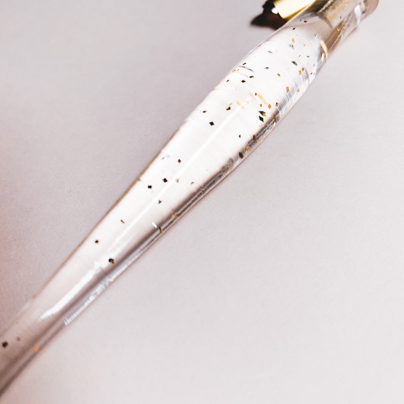 May include: Clear, tapered pen with gold accents and gold glitter flecks suspended within the body. The pen has a gold-colored top and is set against a white background. The pen's design is elegant and decorative.