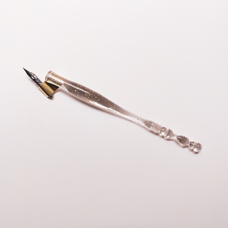 May include: A clear calligraphy pen with a pointed nib and a gold-colored metal nib holder. The pen's handle is transparent with small glitter-like particles embedded within. The handle has a bulbous end and a curved design for comfortable grip.