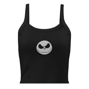 Puede incluir: Un top corto negro sin mangas que presenta un gráfico bordado en blanco centrado de una cara de calavera redonda y sonriente con detalles de boca cosida y ojos huecos en forma de almendra, diseñado con cuello redondo y tirantes anchos.