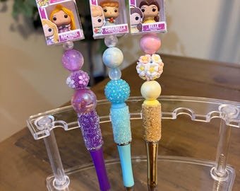 May include: Three decorative pens featuring Disney characters Rapunzel, Cinderella, and Belle. Each pen has a unique design with colorful beads and glitter accents. The pens are displayed in a clear acrylic stand.