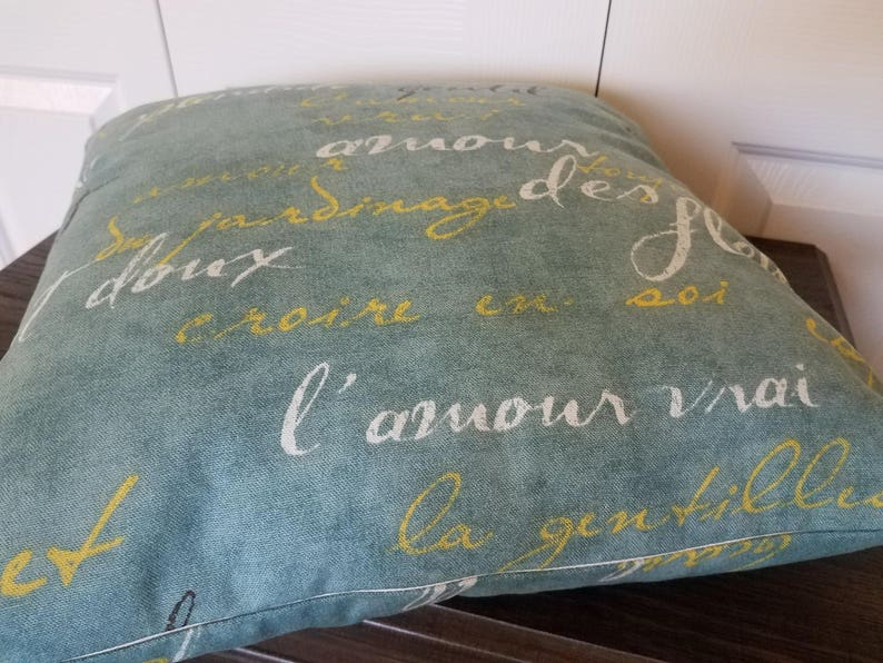 May include: Square decorative pillow with a teal background and French script in white and yellow. The pillow is made of a soft fabric and has a stitched edge. The pillow is placed on a dark wooden surface.