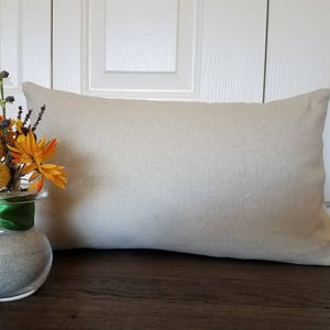 May include: A beige linen pillow with a decorative, natural fiber texture. The pillow is sitting on a wooden surface in front of a white wall.