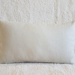 May include: A plain, off-white linen pillow cover. The pillow cover is lying flat on a white fluffy surface.