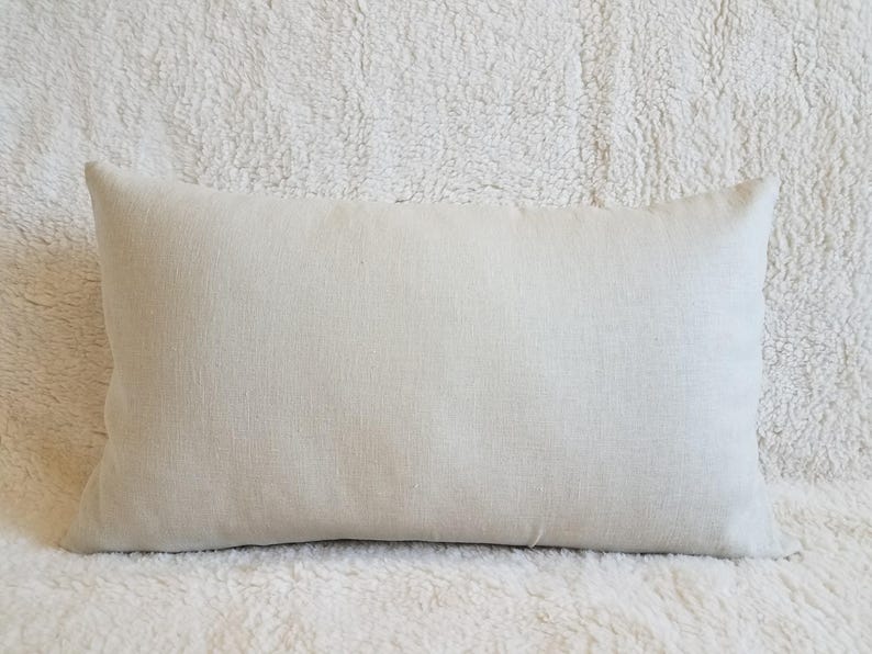 May include: A plain, off-white linen pillow cover. The pillow cover is lying flat on a white fluffy surface.