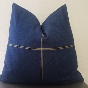 May include: A dark blue denim pillow with yellow stitching. The pillow is square and has a simple design.