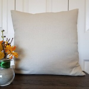 May include: A plain, off-white, linen pillow cover with a subtle texture. The pillow is sitting on a wooden surface. A small vase with yellow and orange flowers is in the foreground.
