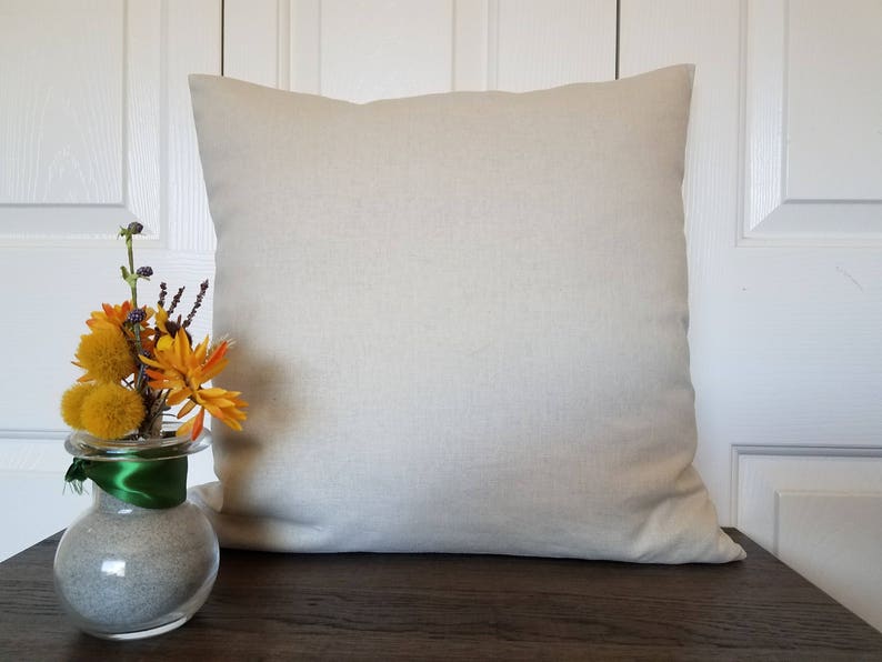 May include: A plain, off-white, linen pillow cover with a subtle texture. The pillow is sitting on a wooden surface. A small vase with yellow and orange flowers is in the foreground.
