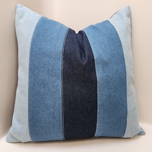 May include: A blue and white striped denim pillow cover. The pillow cover is made of different shades of blue denim fabric sewn together.