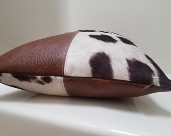 May include: A brown and white cowhide pillow with a brown leather accent. The pillow has a zipper closure.