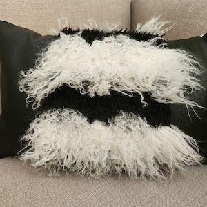 May include: A black and white faux fur pillow with a black leather backing. The pillow is fluffy and has a long, shaggy texture.