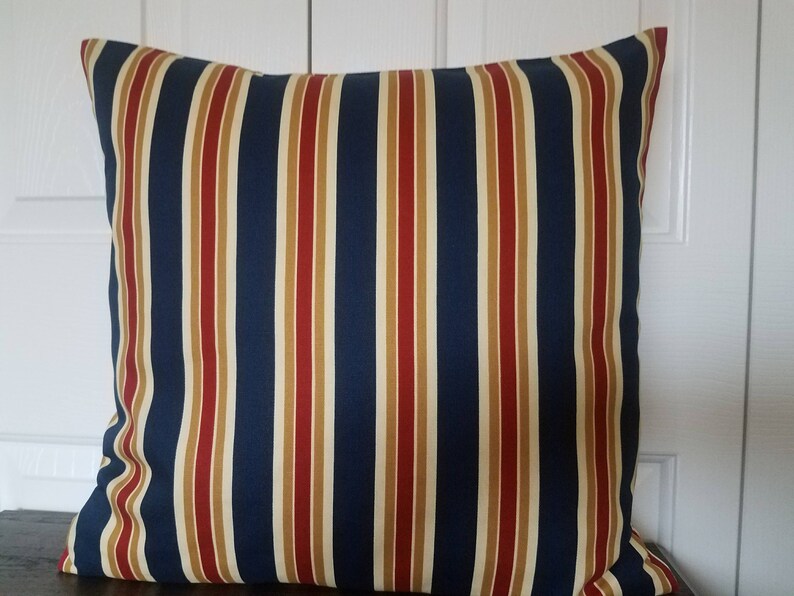 May include: A striped pillow with navy blue, red, and beige stripes. The pillow is made of a soft, woven fabric.