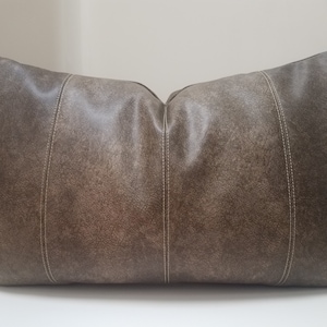 May include: A brown leather pillow with white stitching. The pillow has a textured surface and a rectangular shape.
