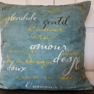 May include: Square teal pillow with French script in white, yellow, and black. The pillow features phrases like "amour" and "gentil" in a decorative font. A home decor item.