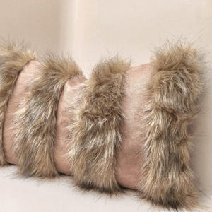 May include: A rectangular decorative pillow with a blush pink center and bands of faux fur trim in shades of brown and tan. The pillow is designed to add texture and warmth to any space.