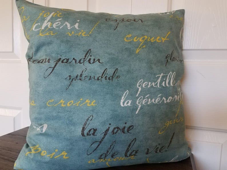 May include: A square teal pillow with French script in yellow and black. The pillow features phrases like "cheri," "jardin," and "la joie." The pillow is on a dark brown surface.