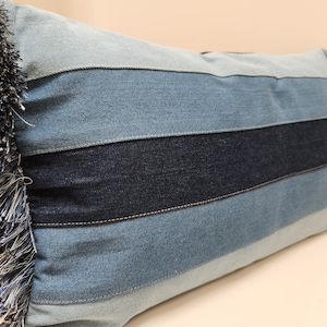 May include: A blue denim pillow with a fringe border. The pillow is made of different shades of blue denim sewn together in horizontal stripes.