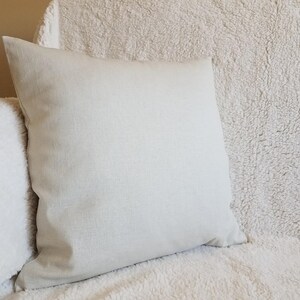 May include: A plain off-white throw pillow with a soft, textured fabric. The pillow is sitting on a white fluffy surface.