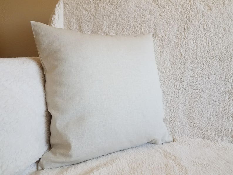 May include: A plain off-white throw pillow with a soft, textured fabric. The pillow is sitting on a white fluffy surface.