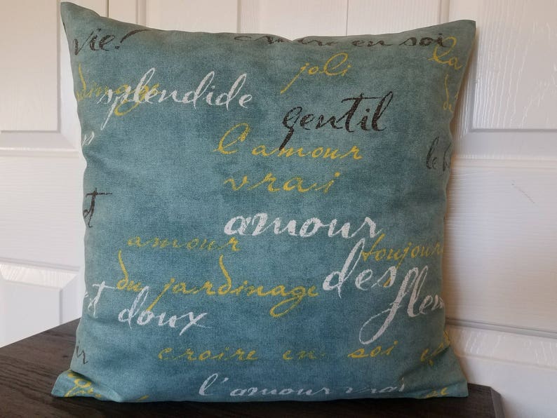 May include: Square teal throw pillow with white, yellow, and black script writing. The pillow has a soft, textured appearance and is placed on a dark brown surface. The script appears to be in French.