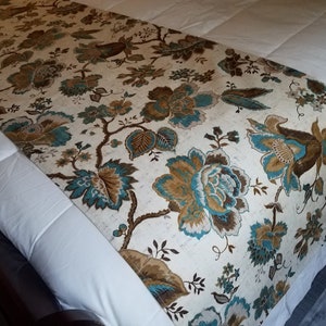 May include: A white bed runner with a floral pattern in shades of brown, teal, and beige. The runner features a large floral design with intricate details.