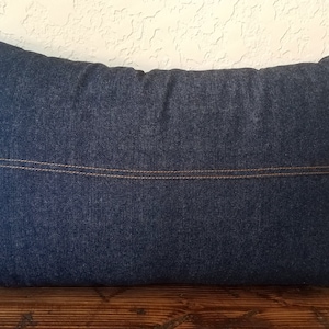 May include: A dark blue denim pillow with a brown stitch detail. The pillow is rectangular and has a smooth, textured surface.