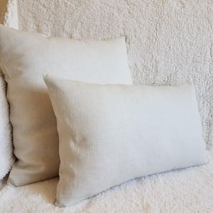 May include: Two off-white rectangular pillows on a white fluffy surface. The pillows are made of a soft, textured fabric.
