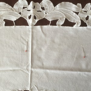 Puede incluir: Primer plano de un textil de lino blanco con un borde bordado floral. El borde presenta dise&ntilde;os de flores y hojas recortadas. Dos alfileres con cabezas rojas est&aacute;n insertados en la tela, lo que sugiere un proyecto de costura.
