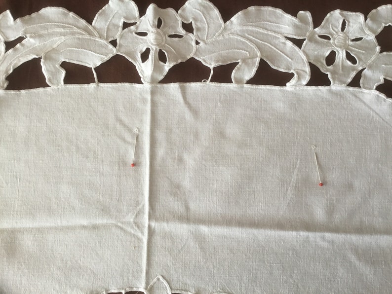 Puede incluir: Primer plano de un textil de lino blanco con un borde bordado floral. El borde presenta dise&ntilde;os de flores y hojas recortadas. Dos alfileres con cabezas rojas est&aacute;n insertados en la tela, lo que sugiere un proyecto de costura.