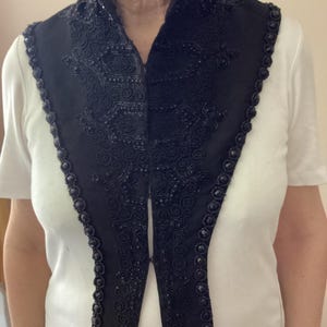 Op de afbeelding: Een zwarte decoratieve kraag met ingewikkeld kralenwerk en kantachtige borduursels. De kraag heeft een hoge, gestructureerde halslijn en een geschulpte rand, wat een formele en elegante uitstraling creëert. De kraag wordt over een wit shirt gedragen.