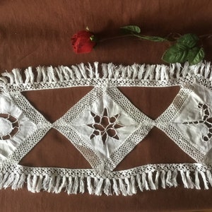 Puede incluir: Camino de mesa de encaje blanco con borde de flecos. El camino de mesa presenta tres recortes en forma de diamante con diseños celestiales: una luna creciente, una estrella y un pájaro estilizado. Una rosa roja y hojas verdes están en el fondo.