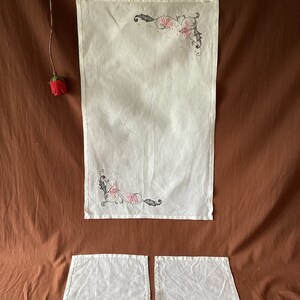 Puede incluir: Un camino de mesa de lino blanco y dos servilletas a juego. El camino de mesa y las servilletas presentan un diseño floral en rosa y gris. Una sola rosa roja está en la esquina superior izquierda. Los artículos están dispuestos sobre una superficie marrón.