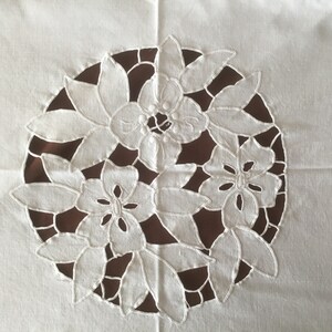 Puede incluir: Dise&ntilde;o floral bordado en blanco sobre fondo de tela blanca. El bordado presenta un patr&oacute;n circular de flores y hojas estilizadas, con un fondo marr&oacute;n visible a trav&eacute;s del dise&ntilde;o recortado. La est&eacute;tica general es delicada y decorativa.