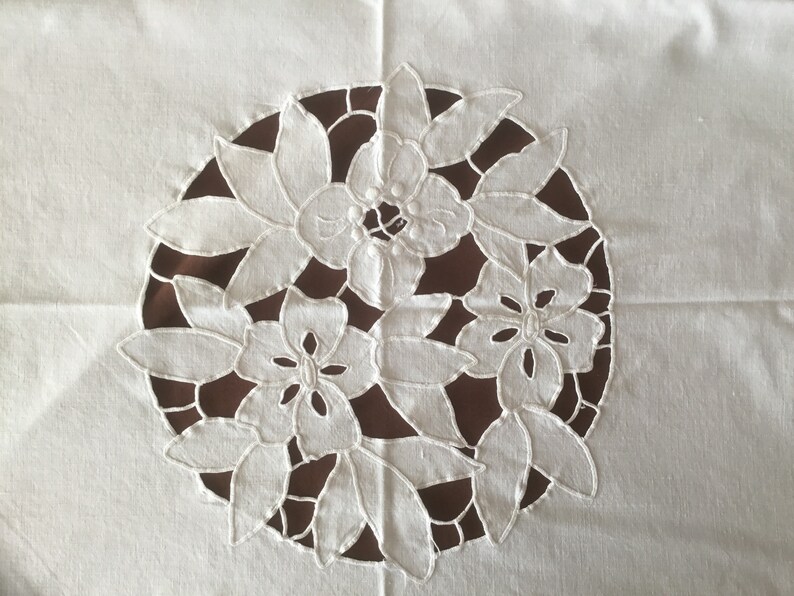 Puede incluir: Dise&ntilde;o floral bordado en blanco sobre fondo de tela blanca. El bordado presenta un patr&oacute;n circular de flores y hojas estilizadas, con un fondo marr&oacute;n visible a trav&eacute;s del dise&ntilde;o recortado. La est&eacute;tica general es delicada y decorativa.