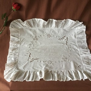 Puede incluir: Funda de almohada rectangular blanca con borde con volantes. El centro presenta diseños de mariposas y flores recortadas. Una rosa roja sobre un tallo está en el fondo.