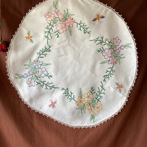 Pode incluir: Uma toalha de mesa redonda bordada branca com uma delicada borda de renda. O tecido apresenta um design floral com flores rosa, azuis e amarelas, juntamente com abelhas bordadas. O bordado é detalhado com caules e folhas verdes.