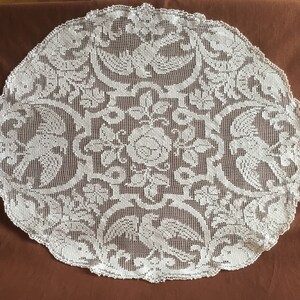 Puede incluir: Un tapete redondo de encaje blanco roto con un detallado diseño floral y de aves. El centro presenta una gran rosa rodeada de hojas y flores más pequeñas. Las aves están simétricamente colocadas alrededor de los bordes, creando una estética vintage.
