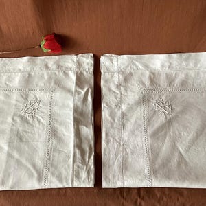 Peut inclure: Deux taies d'oreiller blanches avec des détails brodés et une bordure décorative. Chaque taie d'oreiller présente un monogramme brodé dans le coin. Une rose rouge est placée sur le fond marron. Les taies d'oreiller sont en tissu doux et texturé.