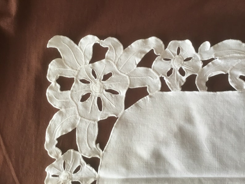 Puede incluir: Primer plano de un dise&ntilde;o floral blanco bordado sobre tela blanca. El dise&ntilde;o presenta flores y hojas caladas, con un patr&oacute;n detallado recortado. El fondo es de color marr&oacute;n liso.