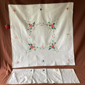 Puede incluir: Un mantel blanco bordado y servilletas a juego. El mantel presenta un diseño floral cuadrado con bordados rosas, azules, amarillos y verdes. Las servilletas tienen un detalle bordado similar.