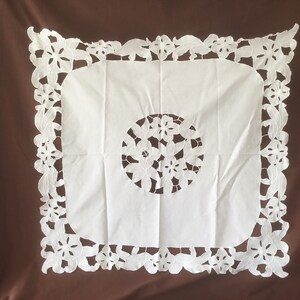Puede incluir: Mantel cuadrado blanco con un dise&ntilde;o floral recortado en el centro y en los bordes. El mantel est&aacute; hecho de una tela blanca suave y est&aacute; sobre un fondo marr&oacute;n.