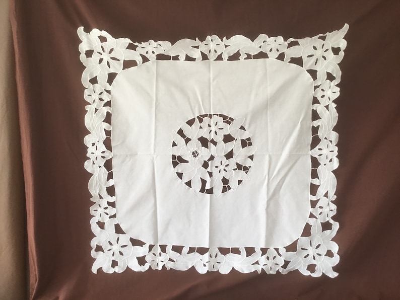 Puede incluir: Mantel cuadrado blanco con un dise&ntilde;o floral recortado en el centro y en los bordes. El mantel est&aacute; hecho de una tela blanca suave y est&aacute; sobre un fondo marr&oacute;n.